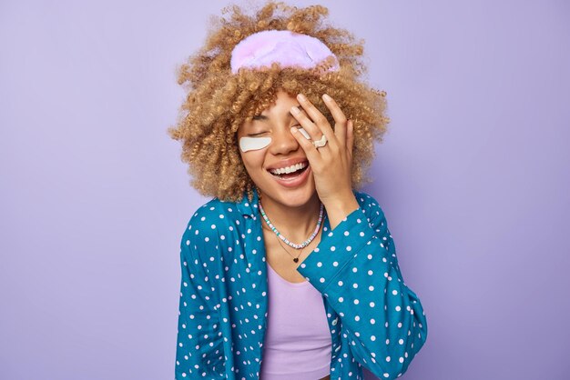 Una mujer feliz y optimista con el pelo rizado hace que la cara de la palma sonría positivamente aplica parches de belleza debajo de los ojos para reducir la hinchazón y usa un antifaz para dormir en la cabeza aislado sobre un fondo morado