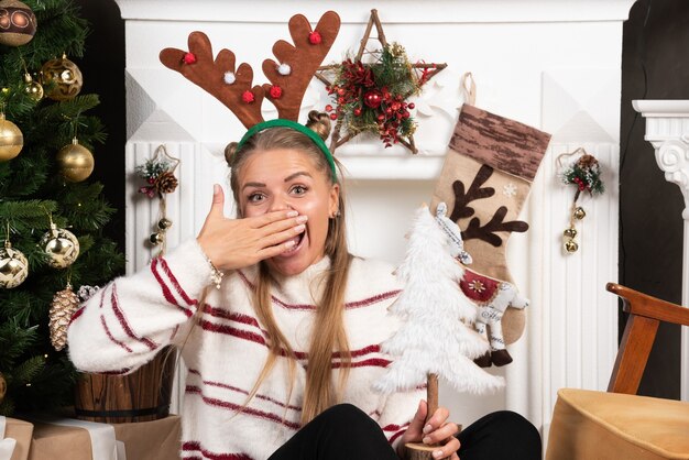 Mujer feliz en orejas de ciervo sosteniendo el árbol de Navidad blanco y tapándose la boca.