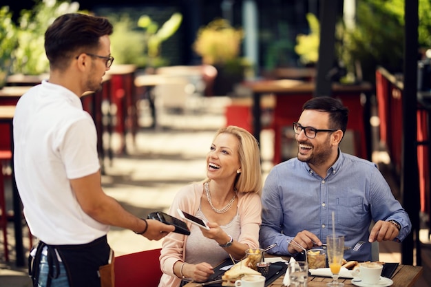 Mujer feliz pagando con su teléfono mediante pago sin contacto mientras almuerza con un novio en un bar