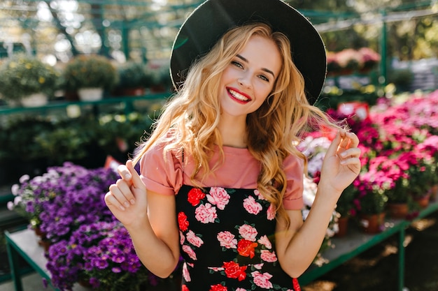 Mujer feliz con peinado rizado posando en invernadero. Espectacular modelo europeo de pie junto a flores.