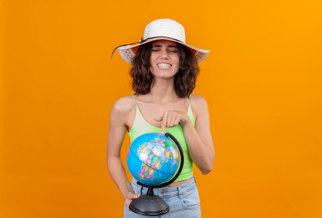 Una mujer feliz con el pelo corto en verde crop top con sombrero para el sol apuntando a un globo con el dedo índice