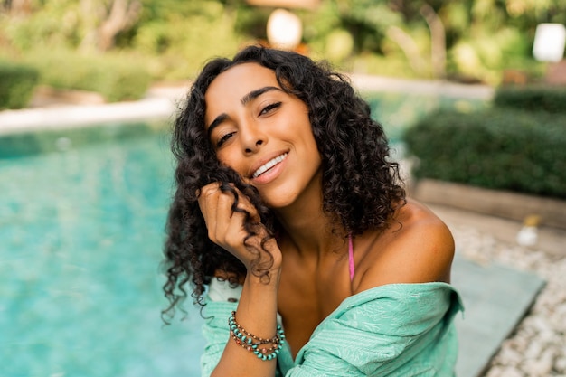 Mujer feliz con pelos ondulados en traje de verano sentado cerca de la piscina Estado de ánimo de vacaciones y viajes Colores brillantes Accesorios de moda