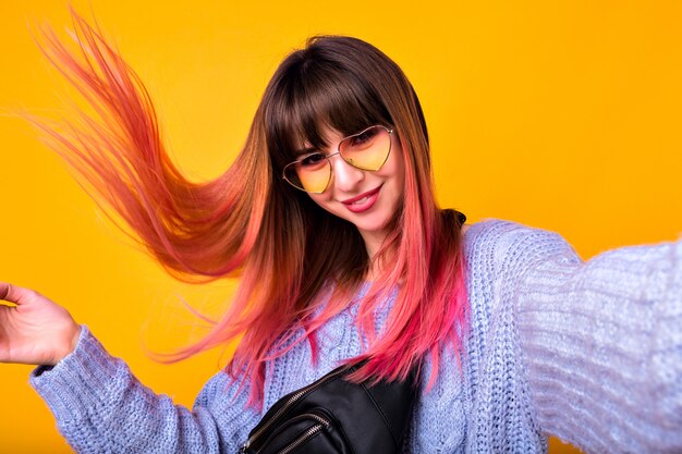 mujer feliz con pelos rosados inusuales haciendo selfie en pared amarilla, suéter elegante y acogedor y gafas de sol vintage con corazón.