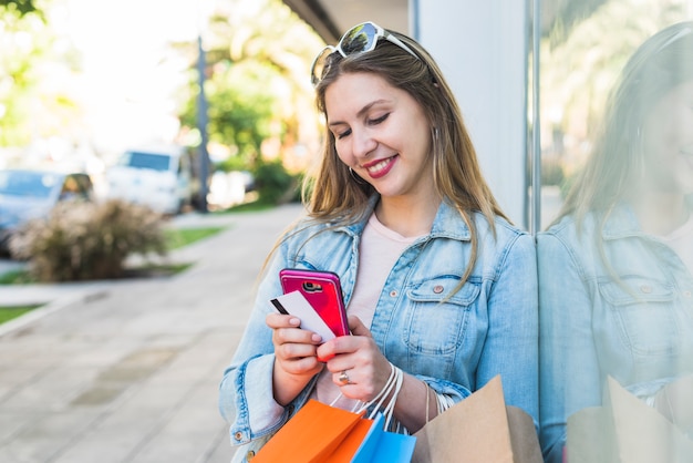 Foto gratuita mujer feliz de pie con bolsas de la compra, teléfono inteligente y tarjeta de crédito fuera