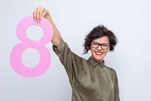 Foto gratuita mujer feliz y positiva con el pelo corto sosteniendo el número ocho de cartón mirando a la cámara sonriendo alegremente celebrando el día internacional de la mujer el 8 de marzo de pie sobre fondo blanco.