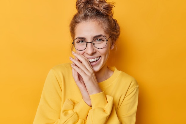 Mujer feliz positiva se ríe con gusto mantiene las manos cerca de la boca expresa auténticas emociones sinceras usa espectáculos y saltador casual aislado sobre fondo amarillo vívido Concepto de felicidad