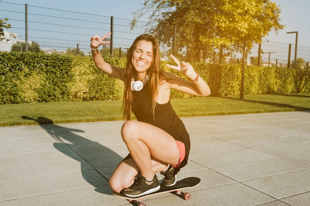 Foto gratuita mujer feliz que gesticula el signo de la paz mientras que anda en monopatín en parque