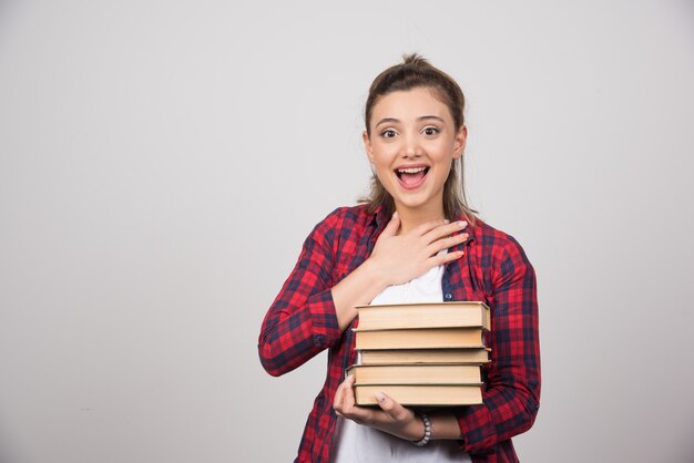 Una mujer feliz que lleva una pila de libros sobre una pared gris.