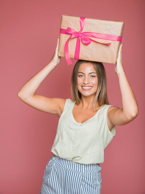 Mujer feliz que lleva el rectángulo de regalo sobre su cabeza contra fondo coloreado