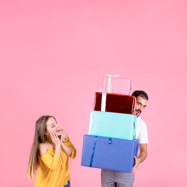 Foto gratuita mujer feliz que mira el hombre que sostiene la pila de cajas de regalo contra el contexto rosado