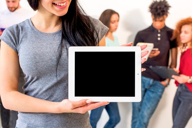 Mujer feliz que muestra la tableta digital de la pantalla en blanco que se coloca delante de sus amigos