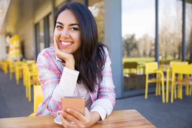 Mujer feliz que se sienta en café de la calle con smartphone y café