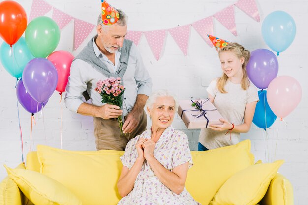 Mujer feliz que se sienta en el sofá delante del marido y de la nieta que sostienen los regalos de cumpleaños
