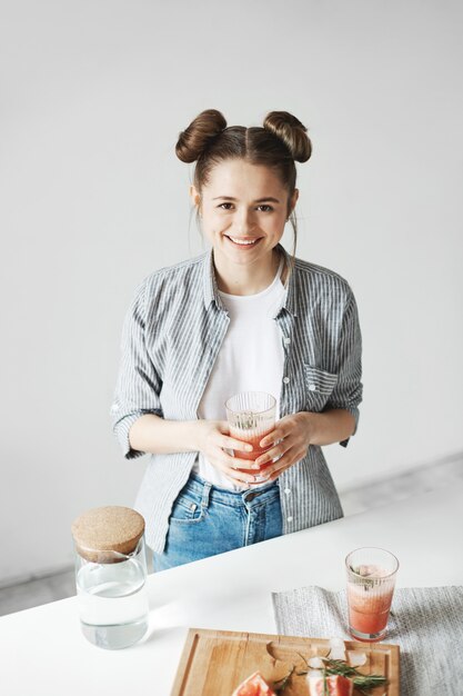 Mujer feliz que sonríe sosteniendo el vidrio con el batido de desintoxicación del pomelo sobre la pared blanca. Comida de dieta saludable.