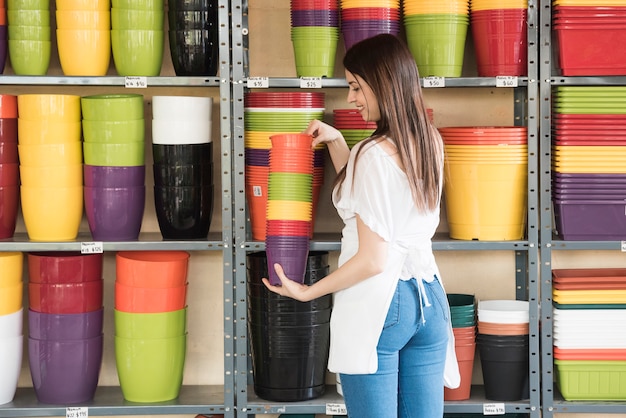 Mujer feliz que sostiene los potes florecientes coloridos delante del estante