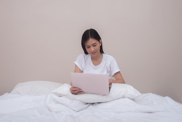 La mujer feliz que usa la computadora portátil que sonríe que se sienta en cama en casa se despertó por la mañana