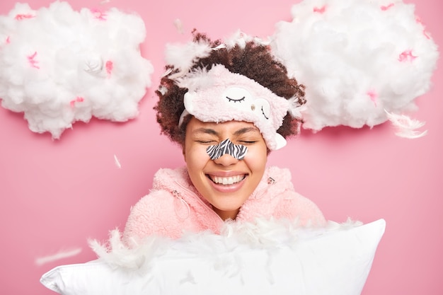La mujer feliz se ríe positivamente mantiene los ojos cerrados aplica un parche en la nariz para eliminar los puntos negros, usa una máscara para dormir y el pijama sostiene una almohada suave rodeada de poses de plumas voladoras en interiores