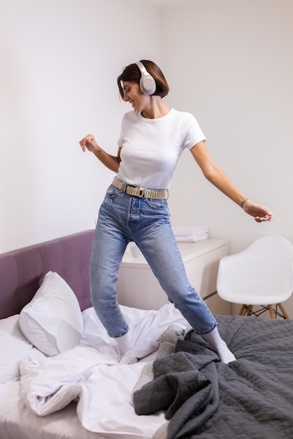 Mujer feliz en ropa casual en casa en el dormitorio escucha música en auriculares, bailando y saltando