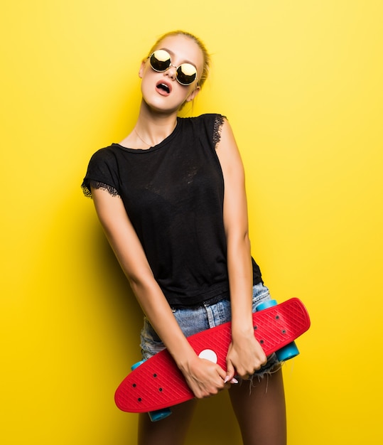 Mujer feliz en ropa de mezclilla y gafas de sol con patineta divirtiéndose y mirando a la cámara sobre fondo amarillo