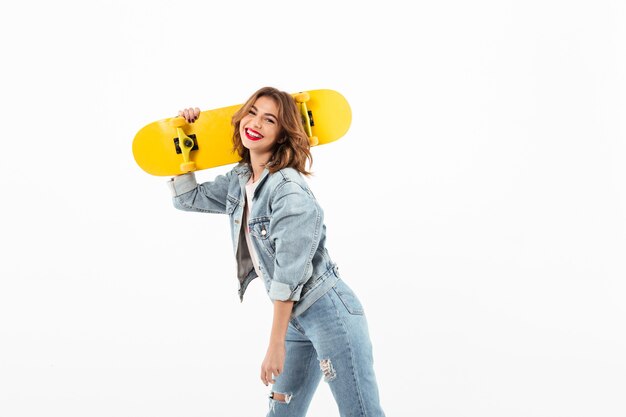 Mujer feliz en ropa de mezclilla posando con patineta sobre pared blanca