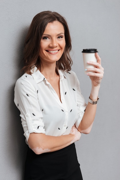 Mujer feliz en ropa de negocios posando cerca de la pared con café en gris