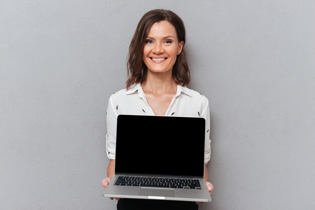 Mujer feliz en ropa de negocios que muestra la pantalla de la computadora portátil en blanco en gris