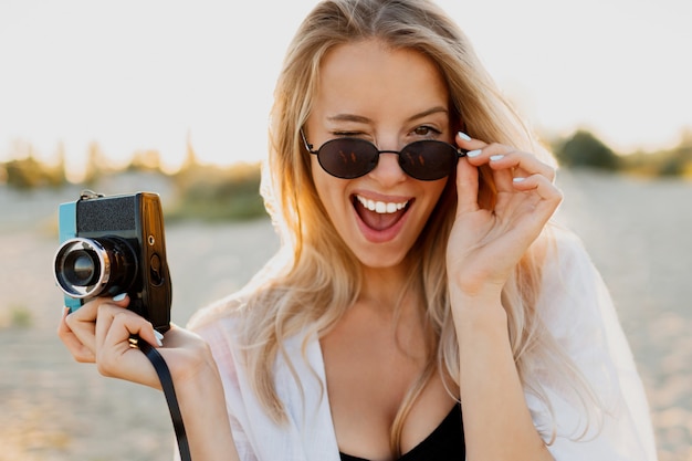 Mujer feliz rubia delgada con cámara retro y divertirse en la cálida playa soleada. Concepto de viaje y vacaciones de verano. Belleza natural, vacaciones en Asia. Gafas de sol de moda, traje blanco.