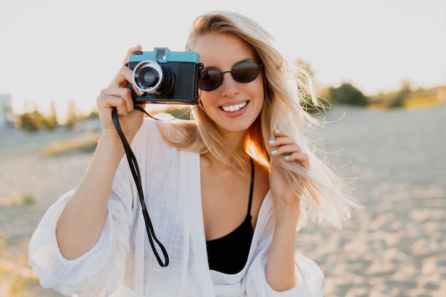 Mujer feliz rubia delgada con cámara retro y divertirse en la cálida playa soleada. Concepto de viaje y vacaciones de verano. Belleza natural, vacaciones en Asia. Gafas de sol de moda, traje blanco.