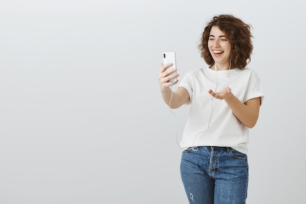 Mujer feliz saliente hablando por videollamada, mensaje de video con amigos mediante teléfono móvil y auriculares