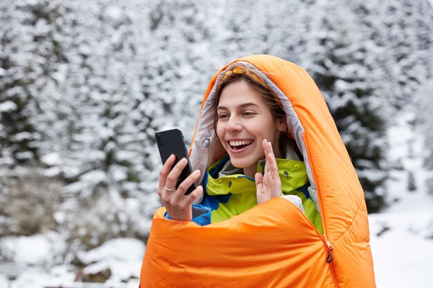 Mujer feliz saluda alegremente a la cámara del teléfono celular, hace videollamadas desde la cima de las montañas cubiertas de nieve