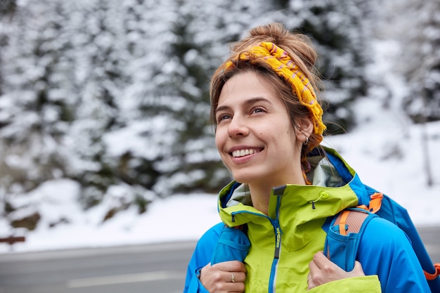 Mujer feliz soñadora con expresión alegre, viste pañuelo amarillo y anorak, lleva mochila