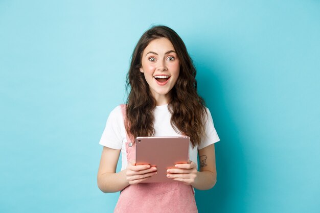 Mujer feliz sorprendida mirando a la cámara asombrada, escuchar noticias increíbles, sosteniendo la tableta en las manos, de pie sobre fondo azul.