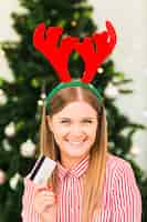 Foto gratuita mujer feliz con tarjeta de plástico en diadema de astas de ciervo cerca de árbol de navidad