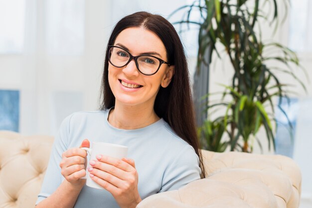 Mujer feliz con la taza de café que se sienta en el sofá