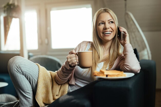 Mujer feliz tomando café y comunicándose por teléfono celular mientras se relaja en el sofá de la sala de estar