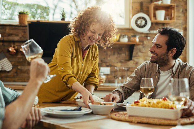 Mujer feliz trayendo comida a la mesa y divirtiéndose con sus amigos durante el almuerzo en casa