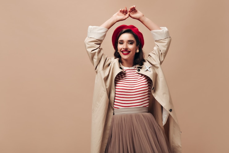Foto gratuito mujer feliz en trinchera y elegante boina bailando sobre fondo beige. encantadora chica con suéter a rayas, falda y abrigo largo sonríe.