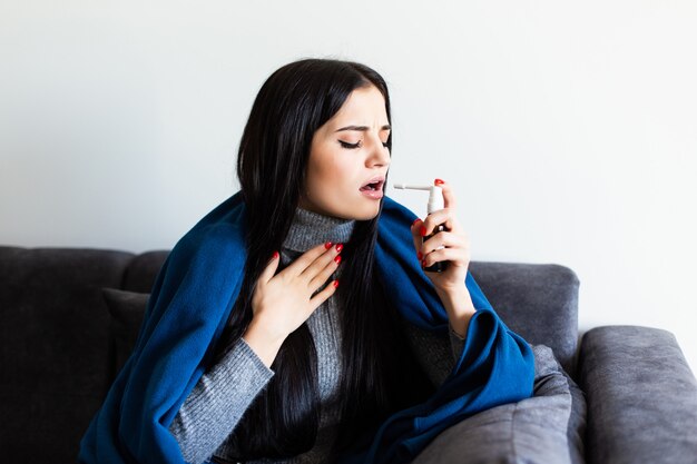 Mujer feliz usando un aerosol analgésico para suavizar la garganta sentado en un sofá en el interior de una casa