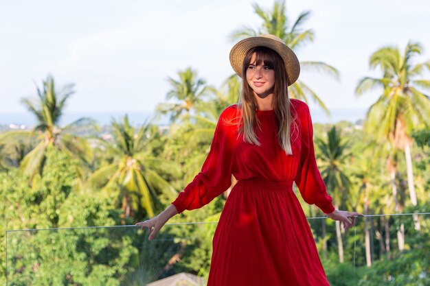 Mujer feliz de vacaciones en vestido rojo de verano y sombrero de paja en el balcón con vista tropical al mar y árboles plam.