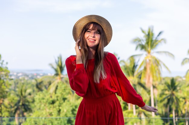 Mujer feliz de vacaciones en vestido rojo de verano y sombrero de paja en el balcón con vista tropical al mar y árboles plam.