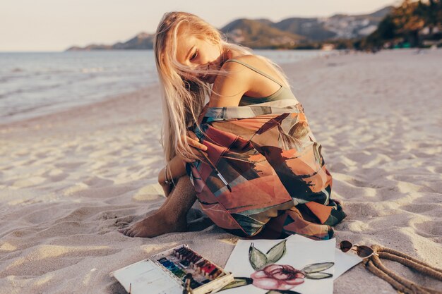 Mujer feliz con ventoso cabello rubio sentado en la arena, mirando su arte de acuarela