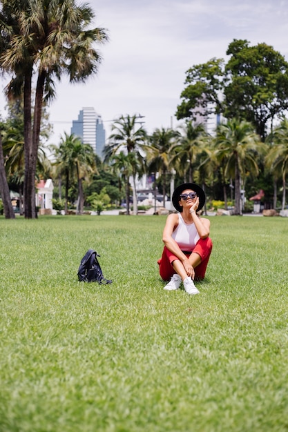 Foto gratuita mujer feliz viajar por bangkok con mochila, disfrutando de un hermoso día soleado en un parque tropical en el campo de hierba verde