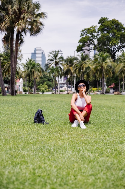 Mujer feliz viajar por Bangkok con mochila, disfrutando de un hermoso día soleado en un parque tropical en el campo de hierba verde