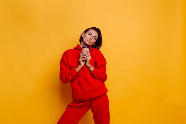 Mujer feliz vistiendo ropa abrigada de invierno, sudadera con capucha y pantalones, se calienta con una taza de café caliente