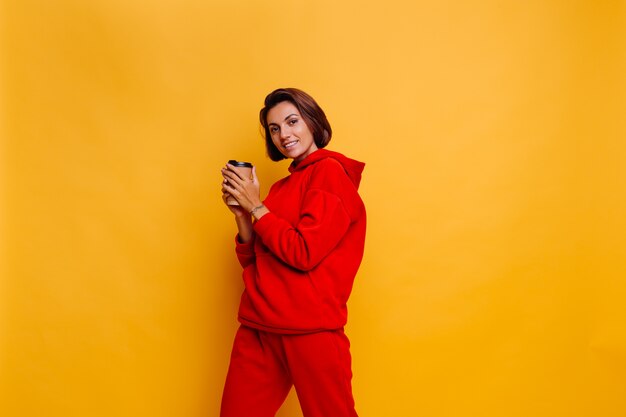 Mujer feliz vistiendo ropa abrigada de invierno, sudadera con capucha y pantalones, se calienta con una taza de café caliente