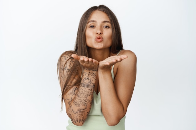 Mujer femenina coqueta con tatuaje, enviando beso al aire en las manos abiertas, lindos labios fruncidos, de pie sobre blanco.
