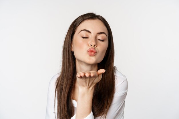 Mujer femenina enviando beso de aire a la cámara, coqueta pose coqueta, besándose, de pie sobre fondo blanco. copia espacio