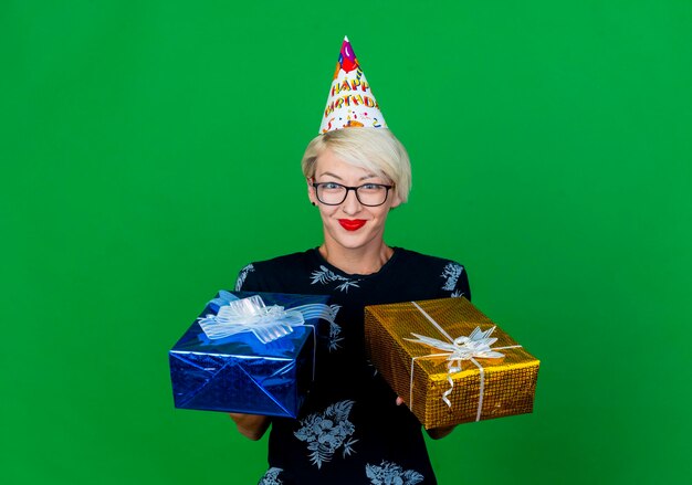 Mujer de fiesta rubia joven complacida con gafas y gorro de cumpleaños sosteniendo cajas de regalo mirando al frente aislado en la pared verde con espacio de copia