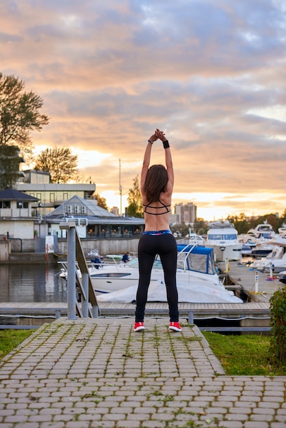 Mujer de fitness deporte descansar después de una intensa carrera nocturna, joven atractiva tomando un descanso después de trotar al aire libre, corredor femenino en ropa deportiva negra mirando la puesta de sol, foto vertical.