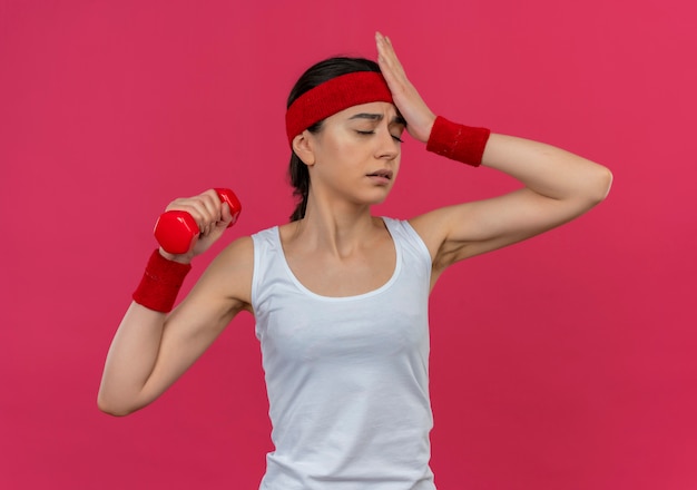Mujer fitness joven molesta en ropa deportiva con diadema sosteniendo mancuernas en la mano levantada mirando confundido con la mano en la cabeza de pie sobre la pared rosa